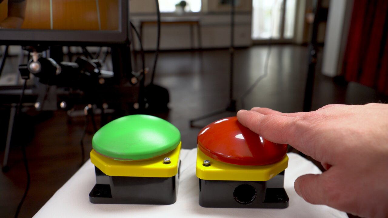 Photo of two big push buttons as commonly used for emergency stops. The right one is red and the left one has been spray painted to be green. A hand is resting above the red button. The rack of the video booth is visible in the background.