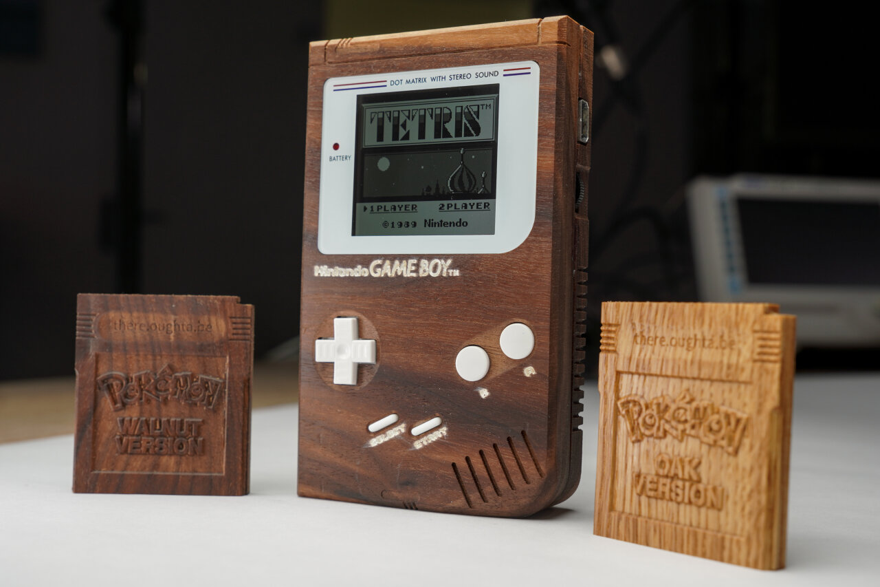 Photo of the wooden Game Boy showing the title screen of Tetris. On its left, there is a wooden cartridge with the label Pokemon Walnut version. On its right, there is a brighter wooden cartridge with the label Pokemon Oak version.