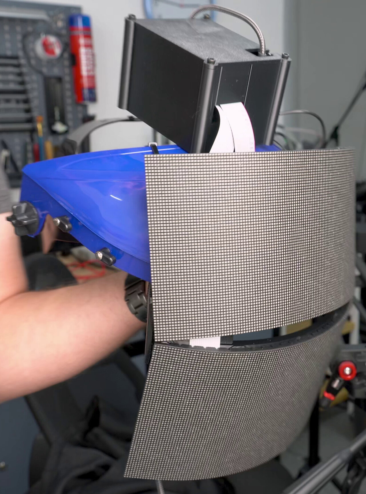Photo of the assembled mask held by a hand. The blue helmet part of a face shield with the two panels dangling from it where the shield would be. An electronics box on top of the helmet is very prominently visible.